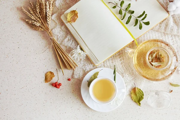 Aesthetic organic linden tea in teapot with cup of tea. Dry fragrance leaves, autumn wheat decorations and mock up notebook top view. Cozy atmosphere home