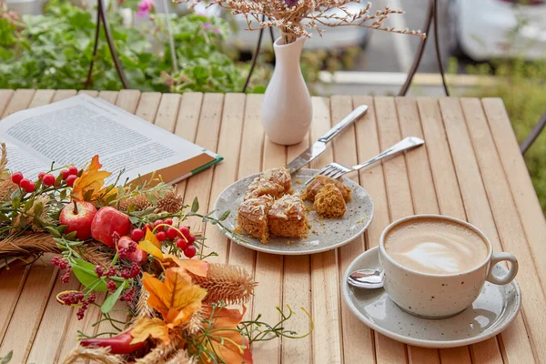 Aesthetic coffee time outside with a book - cutting doughnut and cappuccino among fall decorations on wooden table. Atmospheric coffee time