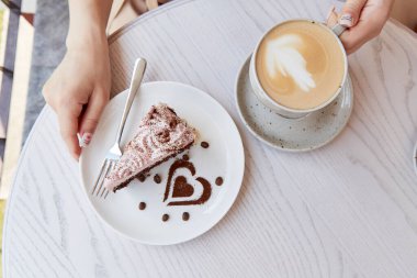 Kadın meyveli çikolatalı kek ve bir fincan kapuçinoyu kafenin terasında, ahşap masada tutuyor. Atmosferik kahve zamanı, kendine zaman ayır. Kalp şeklinde tatlı süslemeleri.