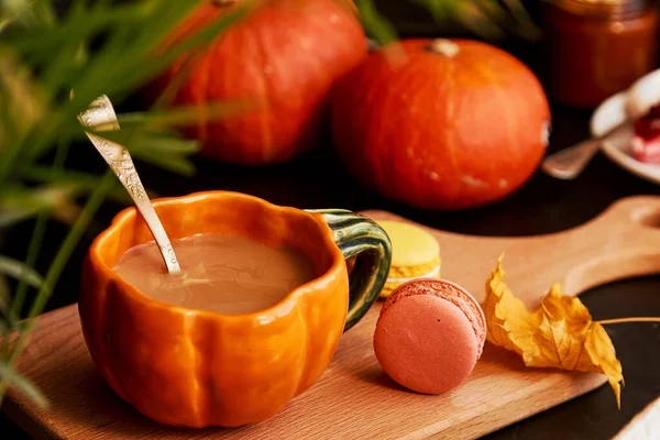 Autumn cup of pumpkin latte in shape of pumpkin, macaroons among pumpkin amd leaves. Atmospheric cozy coffee time.