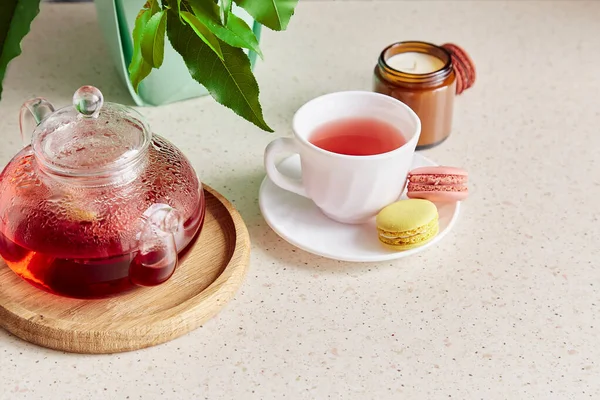 Cozy tea time with cup of tea, teapot and macaroons. Herbal raspberry natural tea in glass teapot. Aesthetic home with candle, dessert. Self care, wellness lifestyle.