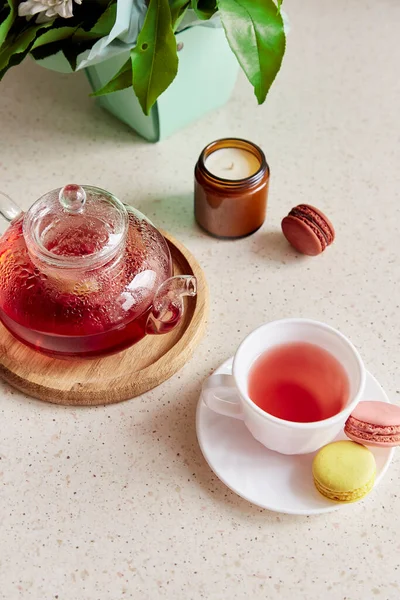 Aesthetic tea time at cozy home. Cup of tea with macaroons. Herbal raspberry natural tea in glass teapot. Cozy home with candle, dessert. Self care, wellness lifestyle.