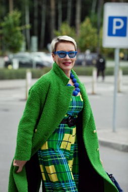 Portrait of fashionable adult woman wearing colorful bright accessories, sunglasses, green stylish coat. Bright fashion model with short blonde hair, street style