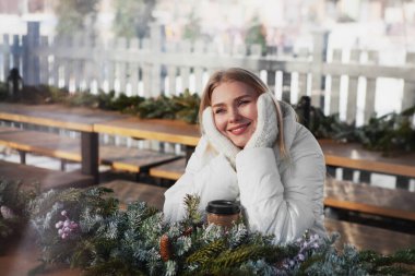 Güzel, gülümseyen sarışın bir kadın ahşap bankta oturur, hayal eder, düşünür, elleriyle tek kullanımlık kağıt bardak ve köknar dallarıyla örülmüş eldivenlerle başını tutar.