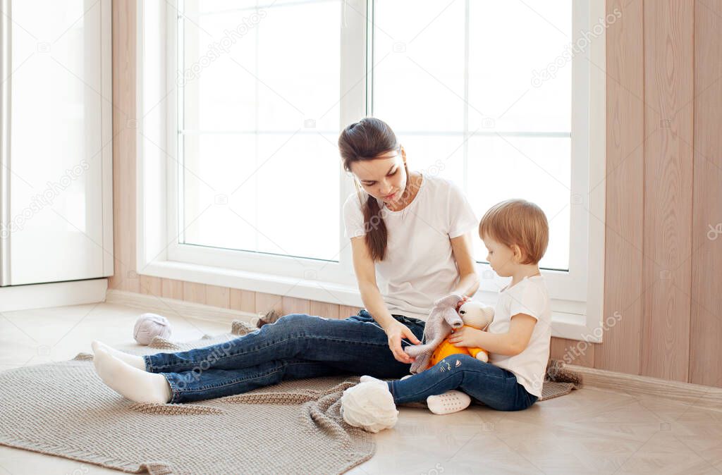 Mom and daughter play dress up soft toy at home next to window in bright modern interior. Mother and small child spend time together, lifestyle