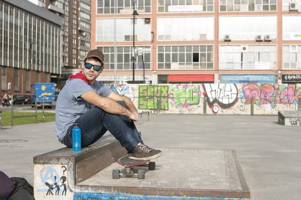 Urban man with blue sunglasses and skateboard sitting on park. — Stock Photo, Image