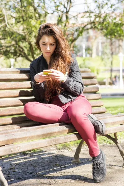 Chica usando el teléfono inteligente y sentado en el banco de madera, al aire libre . —  Fotos de Stock