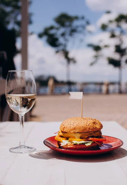 Fresh burger on wooden serving board with onion rings and glass of white wine. Summer beach background