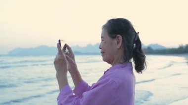 Tatil konsepti 4k Çözünürlük. Sahilde cep telefonuyla oynayan yaşlı bir Asyalı kadın..