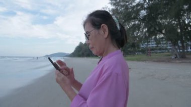 Tatil konsepti 4k Çözünürlük. Sahilde cep telefonuyla oynayan yaşlı bir Asyalı kadın. İnternet iletişimi.