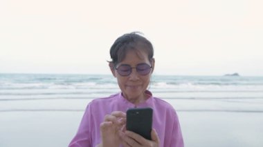 Tatil konsepti 4k Çözünürlük. Sahilde cep telefonuyla oynayan yaşlı bir Asyalı kadın. İnternet iletişimi.