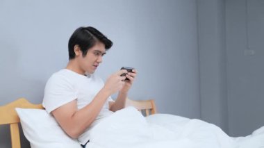 Tatil konsepti 4k Çözünürlük. Asyalı adam yatak odasında mobil oyun oynuyor. online oyun eğlencesi.