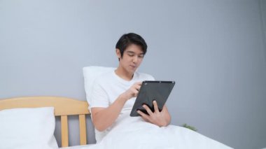Tatil konsepti 4k Çözünürlük. Asyalı adam yatak odasında tablet oynuyor..