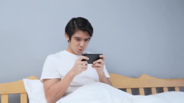 Tatil konsepti 4k Çözünürlük. Asyalı adam yatak odasında mobil oyun oynuyor. online oyun eğlencesi.
