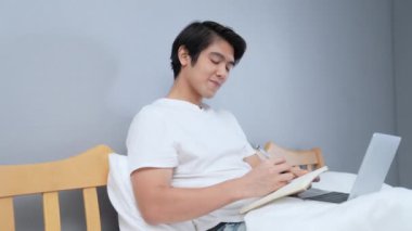 Tatil konsepti 4k Çözünürlük. Yatak odasında çalışan genç bir Asyalı. Not alıyorum..