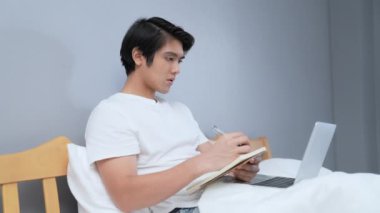 Tatil konsepti 4k Çözünürlük. Yatak odasında çalışan genç bir Asyalı. Not alıyorum..