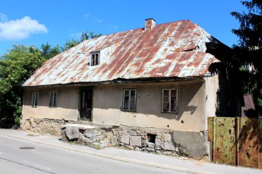 Kırık taş ve beton temeli üzerinde terk edilmiş küçük bir şehir aile evi harap olmuş ahşap çerçeveli tahtadan pencereler ve kaldırımlı yolun yanındaki paslı metal kiremitlerle kaplı kayıp camlarla çevrili hasarlı bir cephe.