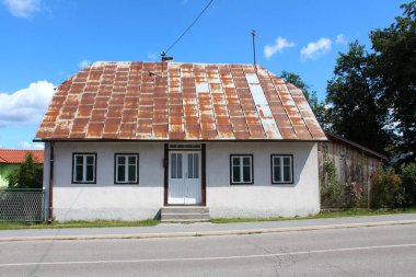 Yenilenmiş ön cephesi ve paslı metal çatı kiremitleri ile kaplı iki beton merdivenin üzerindeki ön çift kapılı girişi ve asfaltlı yolun yanındaki ahşap depolama alanı yüksek ağaçlarla çevrili küçük şehir aile evinin ön manzarası.