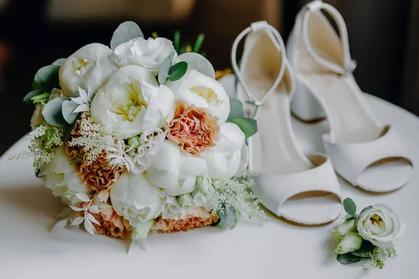 Beyaz bir masada beyaz kadın ayakkabıları topuklu ve bir buket çiçek ve yeşillik — Stok fotoğraf