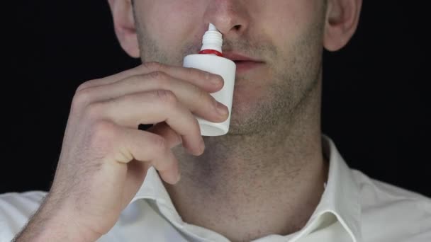 A young man in a white shirt sprays a nasal spray while standing against a black background. Close-up — 비디오