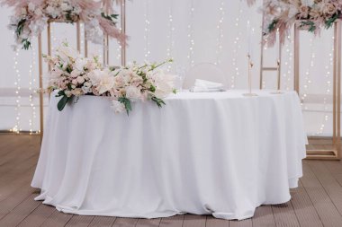  Çiçek ve yeşillik kompozisyonu ile süslenmiş şenlikli beyaz masa masanın üzerinde peçeteler, bardaklar ve mumlarla dolu tabaklar var.