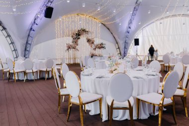 Beyaz masa örtülü ziyafet masaları çadırın altında, masaların üzerinde çiçek aranjmanları var.