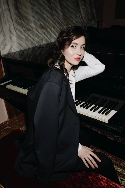 Uma jovem mulher de negócios em um terno preto com cabelo comprido está sentada perto do piano e olhando para a câmera. foto vertical — Fotografia de Stock
