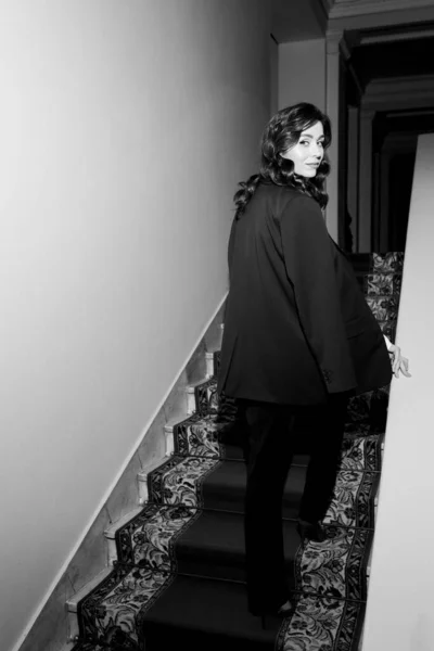 Uma jovem mulher de negócios em um terno preto com cabelo comprido sobe as escadas e olha para a câmera. Foto vertical preto e branco — Fotografia de Stock