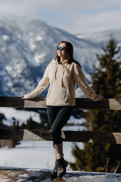  Tarz sahibi, çekici, genç bir kadın terasta açık renk bir ceket, siyah tozluklar ve dağların arka planında duran siyah gözlüklerle duruyor. Yüksek kalite fotoğraf