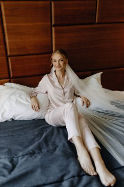 Kahverengi saçlı, peçeli, bej pijamalı, gülümseyen bir kadın kahverengi bir duvara yaslanmış bir yatakta oturuyor. Yüksek kalite fotoğraf