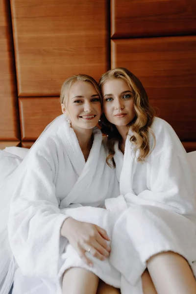 身着白色浴衣的新娘和伴娘坐在床上 近距离看着摄像机 高质量的照片 — 图库照片