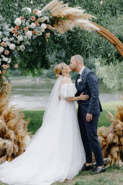 Takım elbiseli genç bir damat ve beyaz uzun elbiseli güzel bir gelin çiçeklerle süslenmiş kemerin yanında nazikçe öpüşüyor. Yüksek kalite fotoğraf
