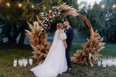 Takım elbiseli genç bir damat ve beyaz uzun elbiseli güzel bir gelin, şefkatle kucaklaşıyorlar, gece ormanlarında çiçeklerle süslenmiş ahşap bir kemerin yanında öpüşüyorlar. Düğün fotoğrafçılığı. 