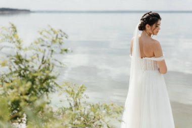 Beyaz gelinlikli gelin sırtı gölde, ağaçların ve çayırların arasında duruyor. Yüksek kalite fotoğraf