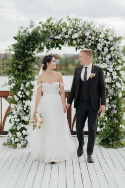 Şirin gülümseyen yeni evliler bir çiçek kemerinin arka planında birbirlerine bakıyorlar. Yüksek kalite fotoğraf