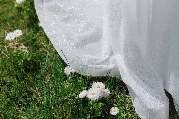 Part of the bride's white shiny dress on green grass with white wildflowers. High quality photo