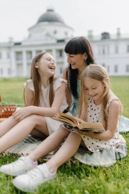  Doğada eğlenen çocuklu genç bir aile. Bir anne ve iki küçük kızı çimlerin üzerinde oturmuş güzel bir parkta gülüyor ve piknik yapıyorlar. Yüksek kalite fotoğraf