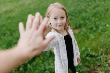  Smiling cute little girl on the background of green grass on a summer day, extending her hand to the photographer for greeting. Copy space. High quality photo