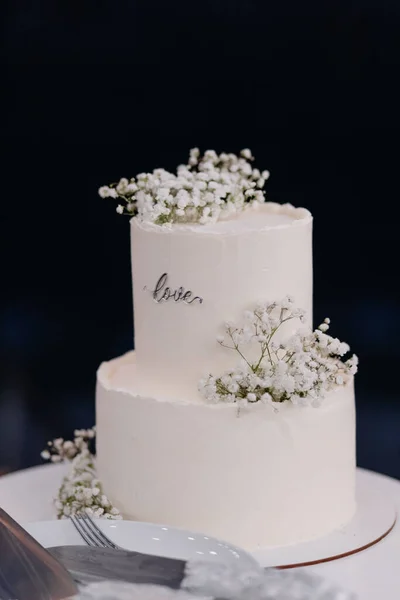 A close up white wedding cake decorated with flowers stands outside on a stand with utensils in the evening. High quality photo