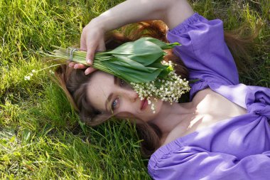 Genç ve güzel bir kız çimlere uzanıyor ve elinde bir buket vadi zambağı tutuyor.