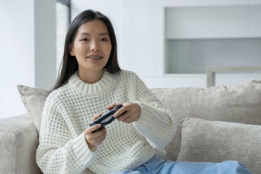 Asyalı bir kadın kanepede oturuyor, elinde bir joystick ve konsolda oyun oynuyor. Kadın joystick ile heyecanlı görünüyor.