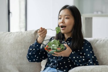 Genç mutlu Asyalı kadın oturma odasındaki kanepede oturmuş sağlıklı salata yiyor..