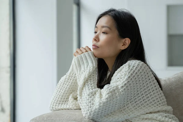 Una joven asiática infeliz sentada en el sofá con una cara triste y mirando por la ventana. — Foto de Stock