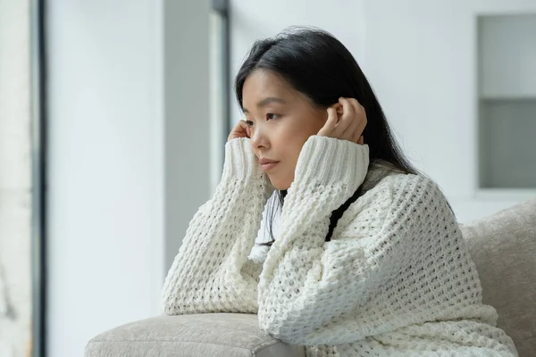Una joven asiática infeliz sentada en el sofá con una cara triste y mirando por la ventana. — Foto de Stock