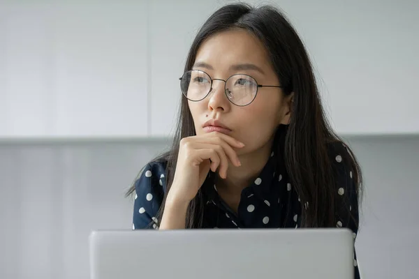 Una mujer de negocios asiática reflexiva con gafas que trabajan con un ordenador portátil mira a la distancia. El estudiante utiliza recursos virtuales de e-learning — Foto de Stock