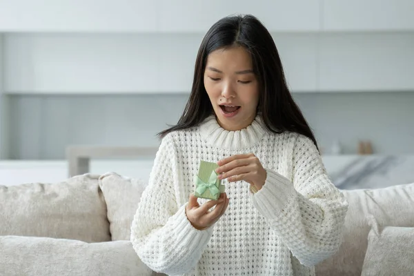 Portret van een gelukkige jonge Aziatische vrouw die een geschenkdoos opent terwijl ze op de bank zit in haar woonkamer — Stockfoto