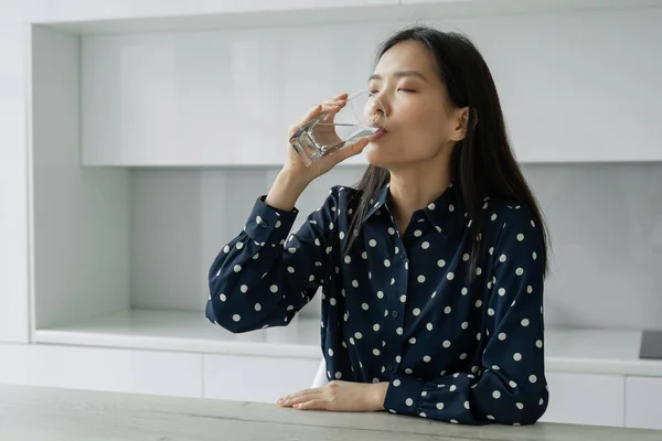 Молодая азиатка пьет воду из стакана, сидящего на кухне. Молодая женщина улыбается, держа стакан воды дома. Концепция образа жизни. — стоковое фото