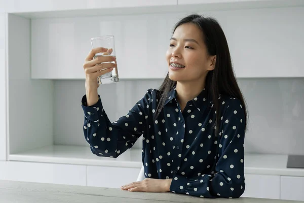 Молодая азиатка пьет воду из стакана, сидящего на кухне. Молодая женщина улыбается, держа стакан воды дома. Концепция образа жизни. — стоковое фото