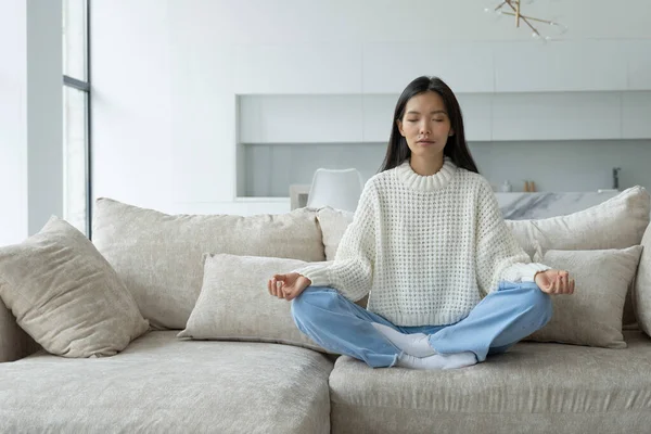 Chica asiática haciendo yoga en la sala de estar sentado en el sofá. Una yogui sentada en la posición de loto. El concepto de yoga y meditación — Foto de Stock