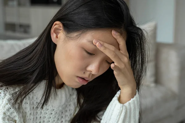 Mano en las sienes de una joven mujer asiática triste sentada en el sofá. Se siente mal por el dolor de cabeza.. — Foto de Stock